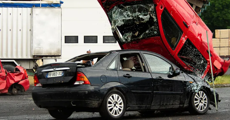 A thrilling car stunt crash with a red car flipped onto a black vehicle in an outdoor setting.