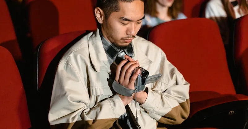 A young man holding a video recorder seated in a movie theater audience.