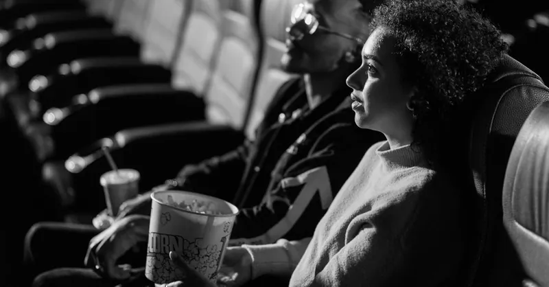 A couple sits together watching a movie in a classic black-and-white cinema setting.