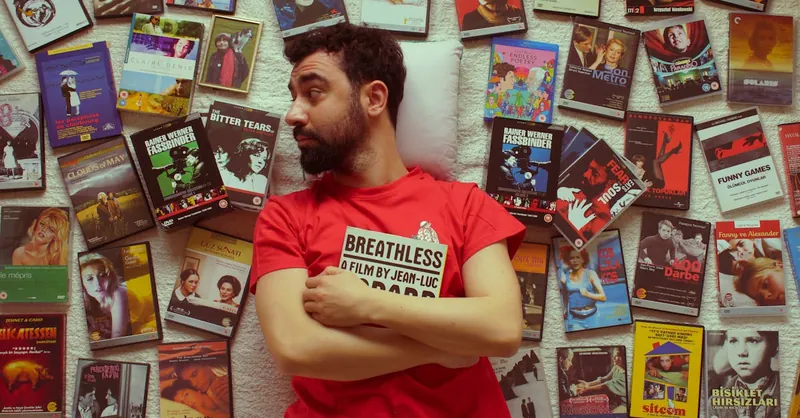 Portrait of a bearded man in a red t-shirt surrounded by vintage movie DVDs, showcasing a love for cinema.